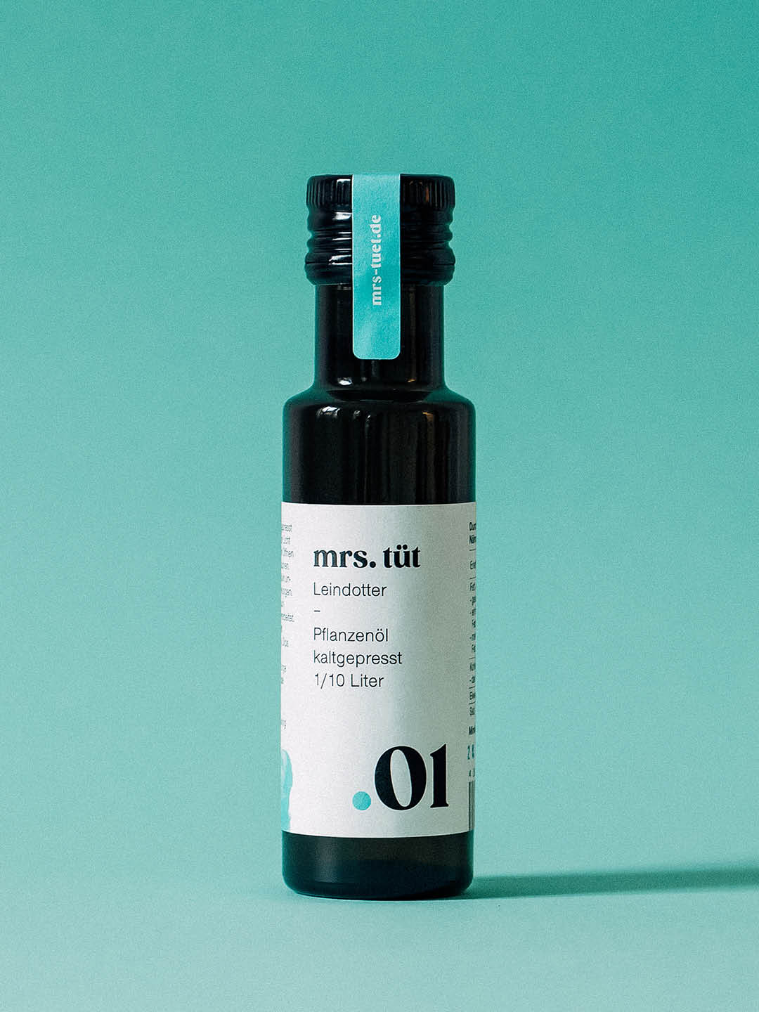 Ölflasche der Marke Mrs. Tüt vor türkisfarbenem Hintergrund mit gestaltetem Etikett